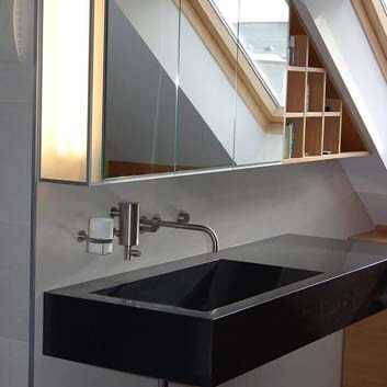 Bathroom with a floating sink countertop, wall-mounted water faucet, and custom-made hanging cabinet with an illuminated side wall, 3 mirror doors, and a shelf under a sloped ceiling