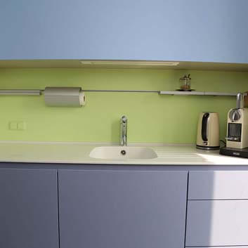 Small office kitchen with molded fronts as handles, blue painted, with white Corian countertop seamlessly integrated with a white Corian sink, Delonghi coffee machine, and green functional backsplash with rail for kitchen towel holder, knife block, and shelves