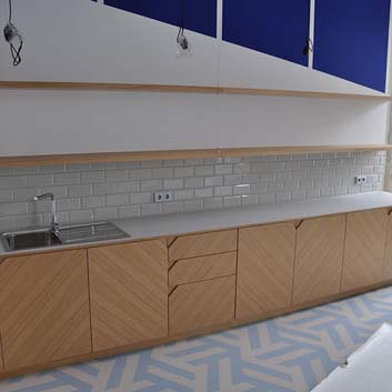 Extended office kitchen with a white countertop, fronts with horizontal wood texture and recessed corners as handles, subway tile backsplash, and open shelves above.
