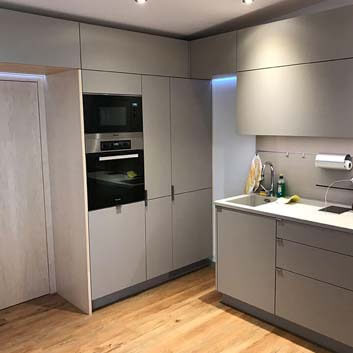 Gray minimalist kitchen with concealed refrigerator, built-in Miele microwave and oven, Corian countertop with seamless kitchen sink, functional splash guard with rail, magnetic knife block.