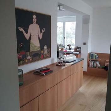 Extension of an open concept kitchen, matching wooden sideboard, with a semi-nude woman cooking who is painting over the sideboard.