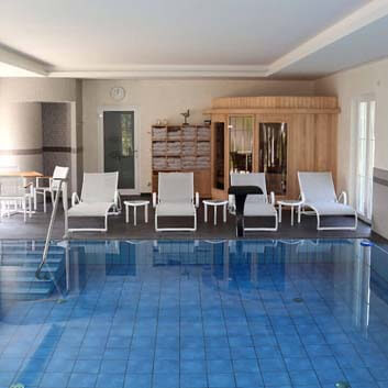 Indoor pool with 4 white loungers and a wooden sauna cabin with glass doors and 2 large glass windows.