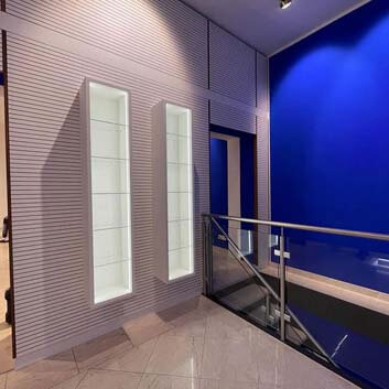 New empty cosmetic salon, one wall with a white ribbed wall-sized panel on a wooden base and two hanging white open glass shelf cabinets, LED lighting on both sides of the inside of the cabinets, the other wall ultramarine blue.