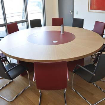 Large round conference table clad in wood like HPL and with a red center table with 3 buttons for covering sockets, with 6 black and 6 red chairs.