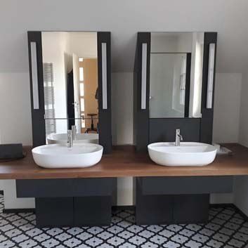 Ein moderner Doppelwaschtisch mit einer dicken Holzarbeitsplatte und zwei rechteckigen, schwarzen vertikalen Paneelen mit Spiegeln und Lampen auf beiden Seiten der Spiegel