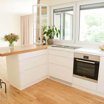 L-förmige weiße Küche mit kürzerer Seite mit einer klobigen Naturholzplatte, die als Frühstückstisch dient, mit einem Barstuhl mit Bora-Herd mit eingebautem Dunstabzug und Bosch-Ofen und einer Vitrine mit Glasregalen auf der Arbeitsplatte