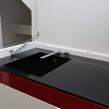 Weißes Küchenelement mit rotem Streifen oben auf den Küchenfronten, schwarze Corian-Arbeitsplatte mit nahtloser Corian-Spüle und abgesenkter Küchenarmatur mit der Möglichkeit, den oberen Deckel zu schließen, um die Küche zu verbergen und das Element wie einen Schrank aussehen zu lassen