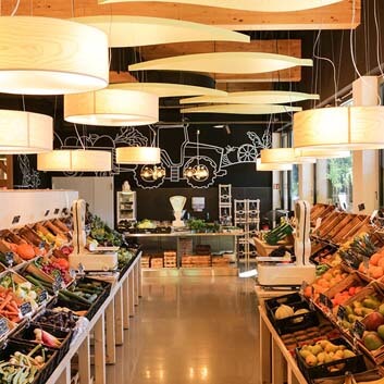 Bio-Supermarkt in Deutschland, hübsches Obst- und Gemüsedisplay in Holzkisten und wiederverwendbaren Plastikkisten auf einem maßgefertigten, geneigten Holzdisplay, Deckenlampen aus Holzfurnier