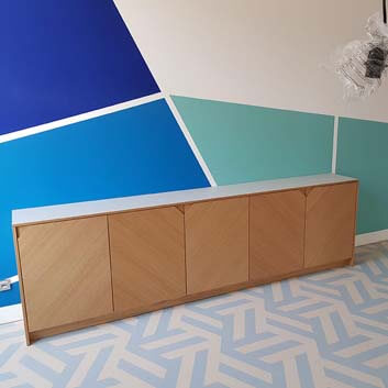 Ein Sideboard aus Holz mit originellen gerillten Ecken an den Fronten, die Griffe, Hintergrundwand und Boden mit blauen und türkisfarbenen Formen bilden