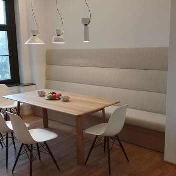 Zum Rand hin ausgedünnter Holztisch, leichte Form, mit 4 elliptischen Holzbeinen, Wand an Wand beige Polsterbank mit überdimensionierter Höhe der Rückenlehne und 5 Vitra Stühlen.