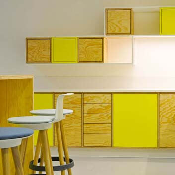 Gemeinsamer Büroraum mit Hängeschränken aus Sperrholz, gelber Möbelplatte und weißem Rahmen.